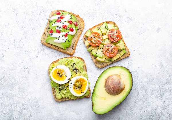 Avocado toasts with egg, tomatoes, seasonings and a half of whole avocado over white stone background. Healthy breakfast avocado sandwiches with different toppings, top view, close-u