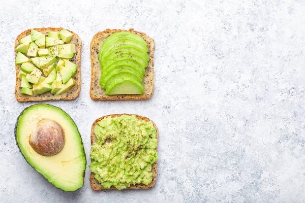Different types of avocado toasts and a half of whole avocado over white stone background, space for text. Ways of making and serving delicious healthy breakfast avocado sandwich, top view, close-u