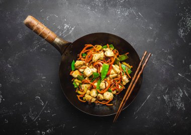 Fry tavuk eski rustik wok tavada sebze, yemek çubukları ile siyah taş zemin üzerine ilave edin, yakın çekim, görünümü top. Geleneksel Asya/Tay yemek