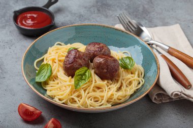 Üzerine köfte ve fesleğen yaprakları konmuş bir tabak spagetti, yanında domates sosu, çatal, kaşık, peçete ve domates dilimleri, sağlıklı ev yemekleri.