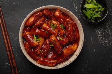 Baharatlı sosla kaynatılmış, yeşil soğan ve susam tohumlarıyla süslenmiş, siyah arka planda servis edilen pirinç kekleri yemek çubukları üst görüşüyle servis edilir. Kore 'nin popüler yemeği.