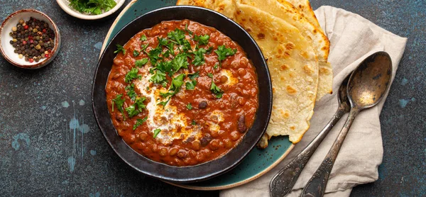 Geleneksel Hint Punjabi yemeği mercimek ve siyah kasede fasulye ile Dal Makhani naan düz ekmek, taze kişniş ve iki kaşık mavi beton kırsal masa manzaralı.