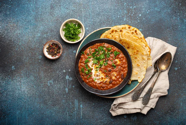 Geleneksel Hint Punjabi yemeği Dal Makhani mercimek ve siyah kasede fasulye ile birlikte naan düz ekmek, taze kişniş ve iki kaşık mavi beton kırsal masa manzaralı. Metin için boşluk.