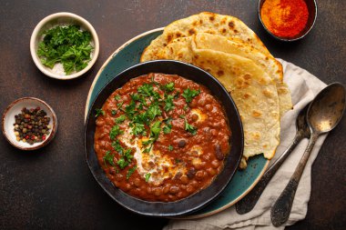 Geleneksel Hint Punjabi yemeği mercimek ve siyah kasede fasulye ile Dal Makhani naan düz ekmek, taze kişniş ve kahverengi beton kırsal masa manzaralı iki kaşık ile servis edilir..