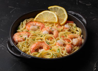 Siyah dökme demir tavada servis edilen karidesli makarna taze limon dilimleri ve siyah kırsal arka planda maydanoz açı görüşüyle süslenmiş.