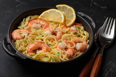 Siyah dökme demir tavada servis edilen karidesli makarna taze limon dilimleri ve siyah kırsal arka planda maydanoz açı görüşüyle süslenmiş.