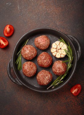 Çiğ köfteler sarımsak ve biberiyeyle tatlandırılmış, dökme demir bir tavada pişirilmeye hazır, koyu kahverengi bir masada, kırmızı kareli peçete ve vişneli domatesle.