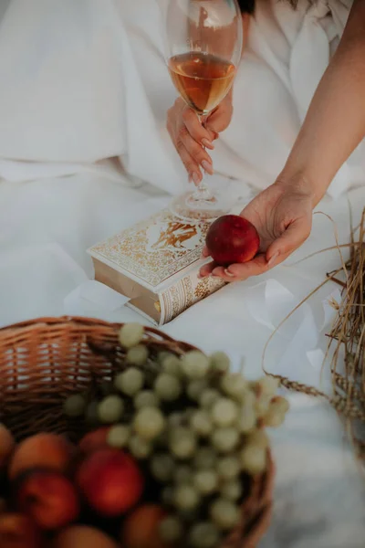 Woman at a picnic. All in white drinking wine, reading a book on a white bedspread. The girl on the picnic gives it to the field. The girl drinks wine and eats fruit. The woman holds a peach in her palm.