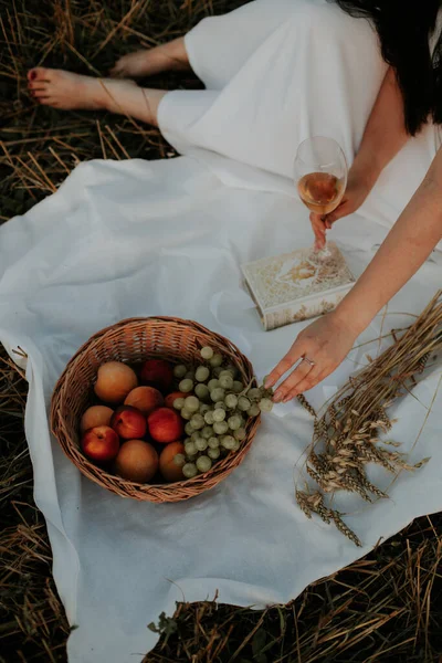 Woman at a picnic. All in white drinking wine, reading a book on a white bedspread. The girl on the picnic gives it to the field. The girl drinks wine and eats fruit.