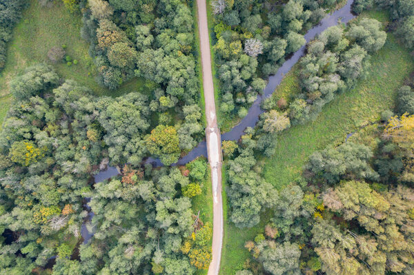 Aerial top down view of gravel road and bridge over winding river flowing through green forest