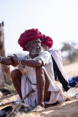 Puşkar, Rajasthan, Hindistan - 10 Kasım 2024: Geleneksel beyaz giysili rajasthani erkek portresi çöl panayırında renkli türbanlı deve fuarı sırasında develeri takas etmek için.