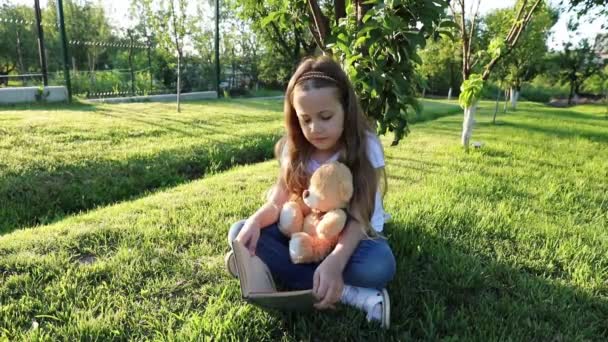 Lány Szabadban Olvas Kis Plüssmacijának Kislány Játékokkal Játszik Zöld Gyepen — Stock videók