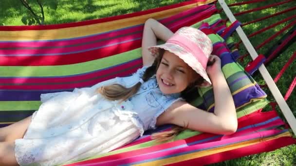 公園の路上でハンモックに寝そべっている少女 明るいハンモックの上に横たわる帽子の小さな甘い女の子 公園の女の子庭で — ストック動画