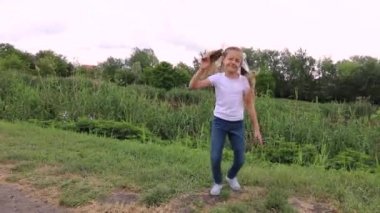 Kot pantolonlu, beyaz tişörtlü, 7-10 yaşlarında komik sarışın kız çocuğu. Beyaz tişörtlü ve kot pantolonlu çocuk zıplıyor. Küçük kız yeşil bir parkta dans ediyor