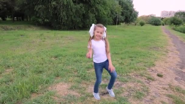 少し楽しいブロンドの子供の女の子7 10デニムで歳 白いTシャツポーズ 白いTシャツとジーンズの子供がジャンプします 緑の公園で踊る少女たち — ストック動画