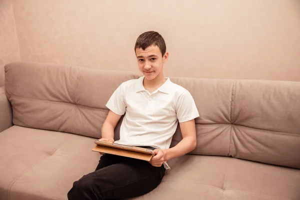 白いTシャツを着た10代の男の子がラップトップでソファに座っています 家庭教育子供の自己教育 — ストック写真