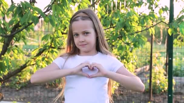 髪の長い可愛い女の子がハートのシンボルを見せてくれます 公園の赤ん坊だ 緑の庭の森の少女 — ストック動画