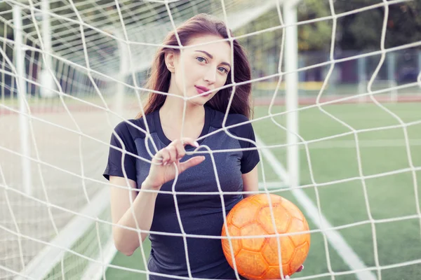 Young woman with a ball in hands. She looks through the net of the football goal.