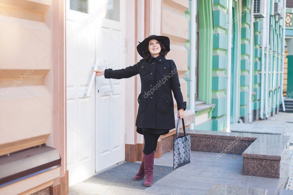 Autumn in the city. Full young woman in coat and hat standing at the white door.