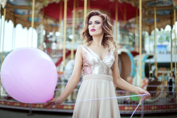 美丽的女人穿着晚礼服站在旋转木马附近 拿着一个气球 — 图库照片