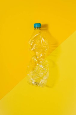 Sarı arka planda mavi kapaklı şeffaf ve ezilmiş plastik bir şişe ile dikey renk görüntüsü. Geri dönüşüm ve çevre kavramı.
