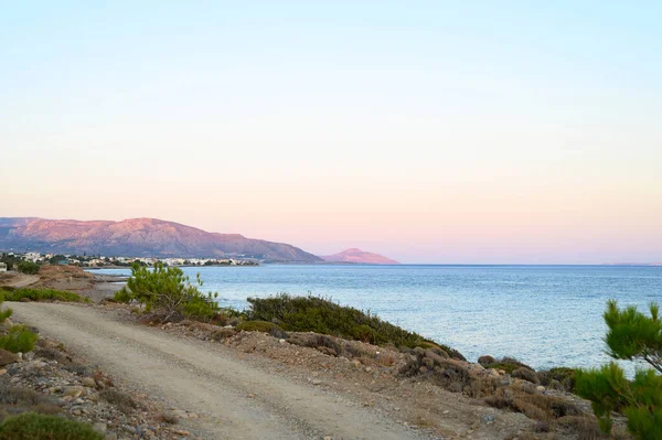 seascape at dusk. a dirt road, a city in the distance and mountains with a gentle pink sunset and a beautiful blue sea