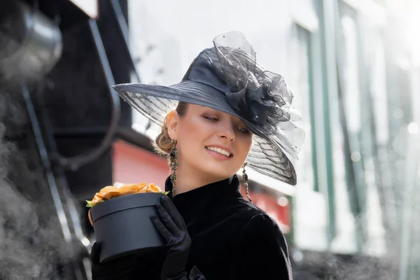 Steampunk girl in a black dress and hat near an old steam locomotive and large iron wheels. Blond beauty. Vintage portrait of the last century, retro journey.