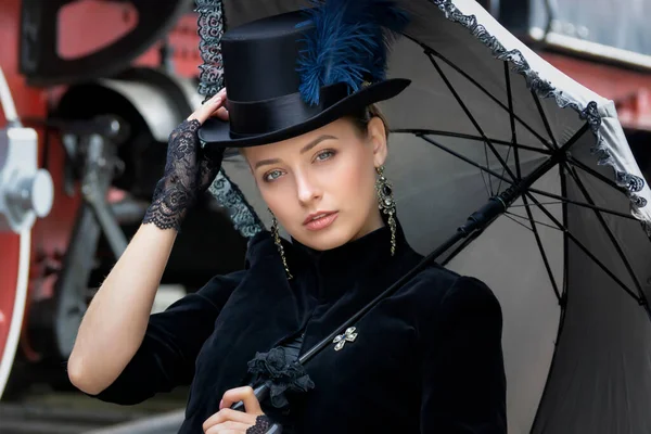 Steampunk girl in a black dress and hat near an old steam locomotive and large iron wheels. Blond beauty. Vintage portrait of the last century, retro journey.