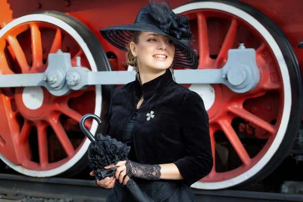 Steampunk girl in a black dress and hat near an old steam locomotive and large iron wheels. Blond beauty. Vintage portrait of the last century, retro journey.