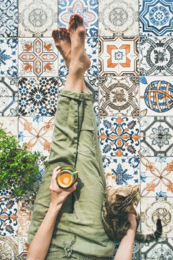 Lazy morning on terrace. Womans legs in cosy linen pants, plant, cat and cup of coffee in hand over colorful moroccan tile floor clipart