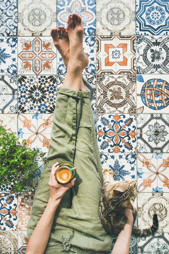 Lazy morning on terrace. Womans legs in cosy linen pants, plant, cat and cup of coffee in hand over colorful moroccan tile floor
