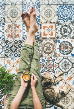 Womans legs in cosy linen pants, plant, cat and cup of coffee in hand over colorful moroccan tile floor clipart