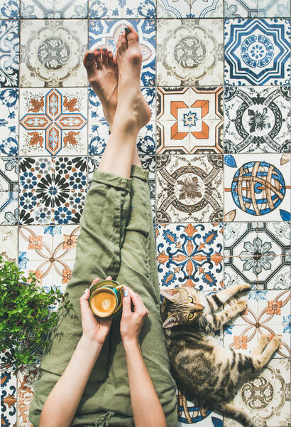 Womans legs in cosy linen pants, plant, cat and cup of coffee in hand over colorful moroccan tile floor
