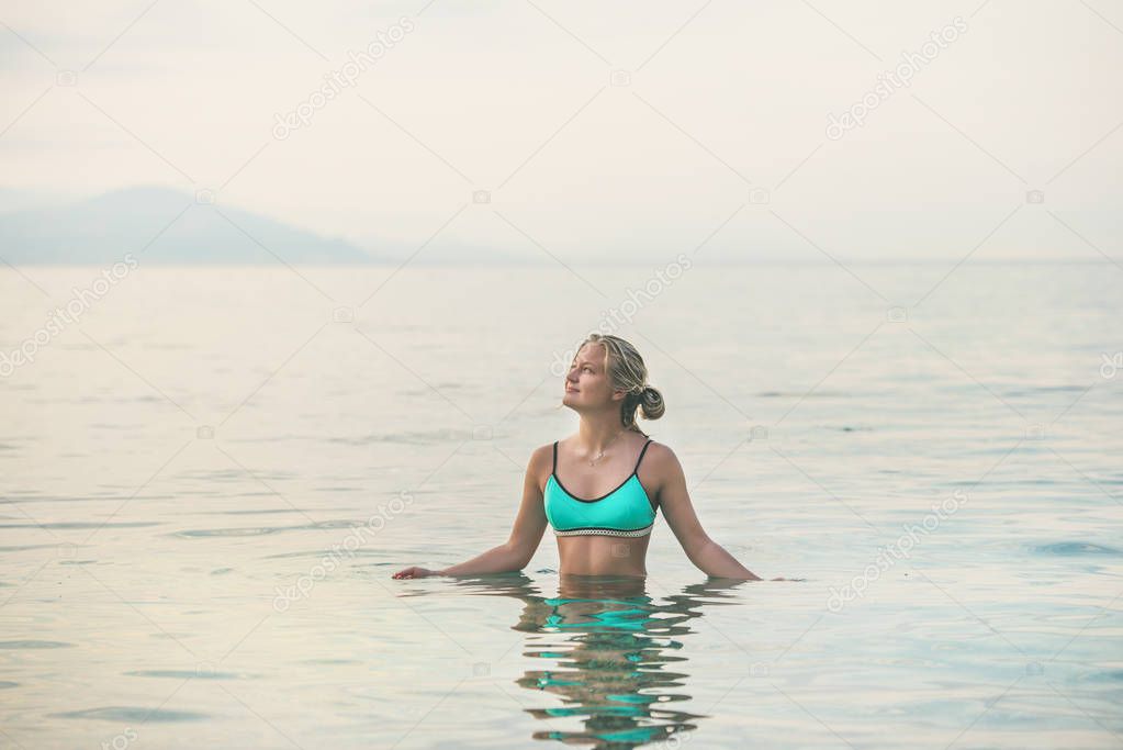 Young beautiful slavian woman tourist wearing turquoise swimsuit standing in still sea waters at beach near ancient fortress wall and shipyard in city center of Alanya, Mediterranean Turkey