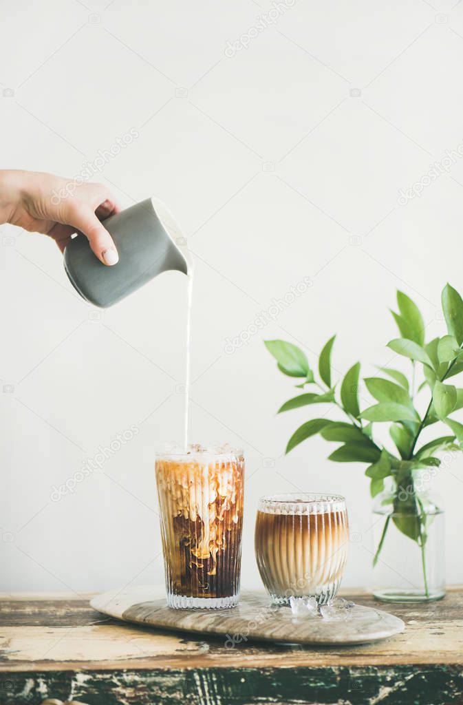 Iced coffee in tall glasses with milk poured over from pitcher by hand, white wall and green plant branches in vase at background