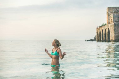 Genç güzel slavian kadın turist turkuaz bikini rahatlatıcı antik kale duvarı ve Alanya şehir merkezine, Akdeniz Türkiye tersanelerinde yakınındaki plajda denize giyiyor