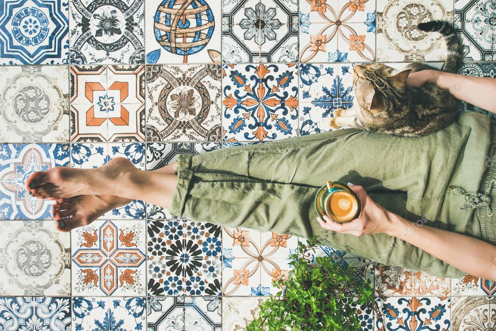 Womans legs in cosy linen pants, plant, cat and cup of coffee in hand over colorful moroccan tile floor