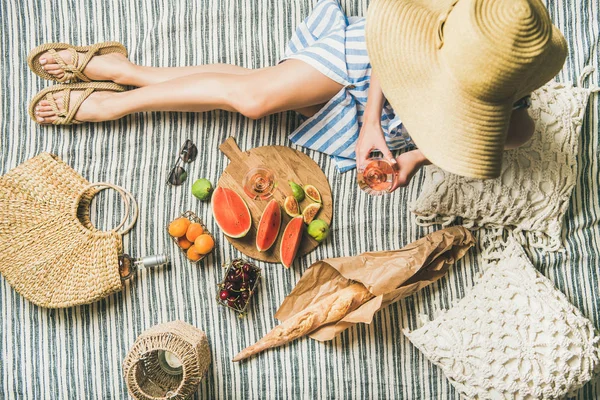 Summer picnic setting. Woman in linen striped dress and straw sunhat sitting with glass of rose wine in hand, fresh fruit and baguette on blanket