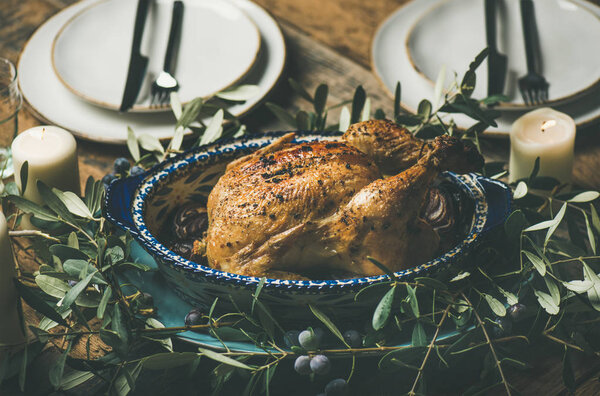 Whole roasted chicken in tray for Christmas eve celebration decorated with olive tree branch, plates and candles over rustic wooden table background
