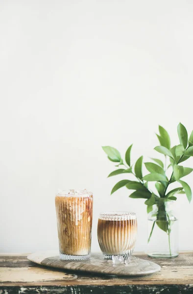 Iced coffee in tall glasses with milk on board, white wall and green plant branches at background