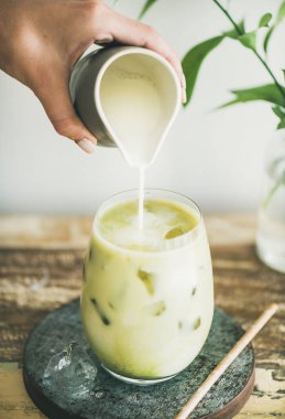 Buzlu matcha latte ile Hindistan cevizi sütü kadının el, beyaz arka plan, duvar ve bitki şubeleri sürahi dökme cam içki