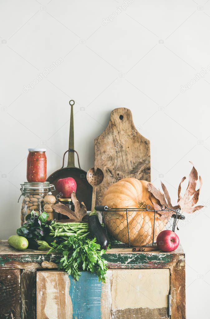 Autumn seasonal food ingredients and kitchen utensils. Vegetables, pumpkin, red apples, canned food, fallen leaves over rustic wooden chest of cupboard