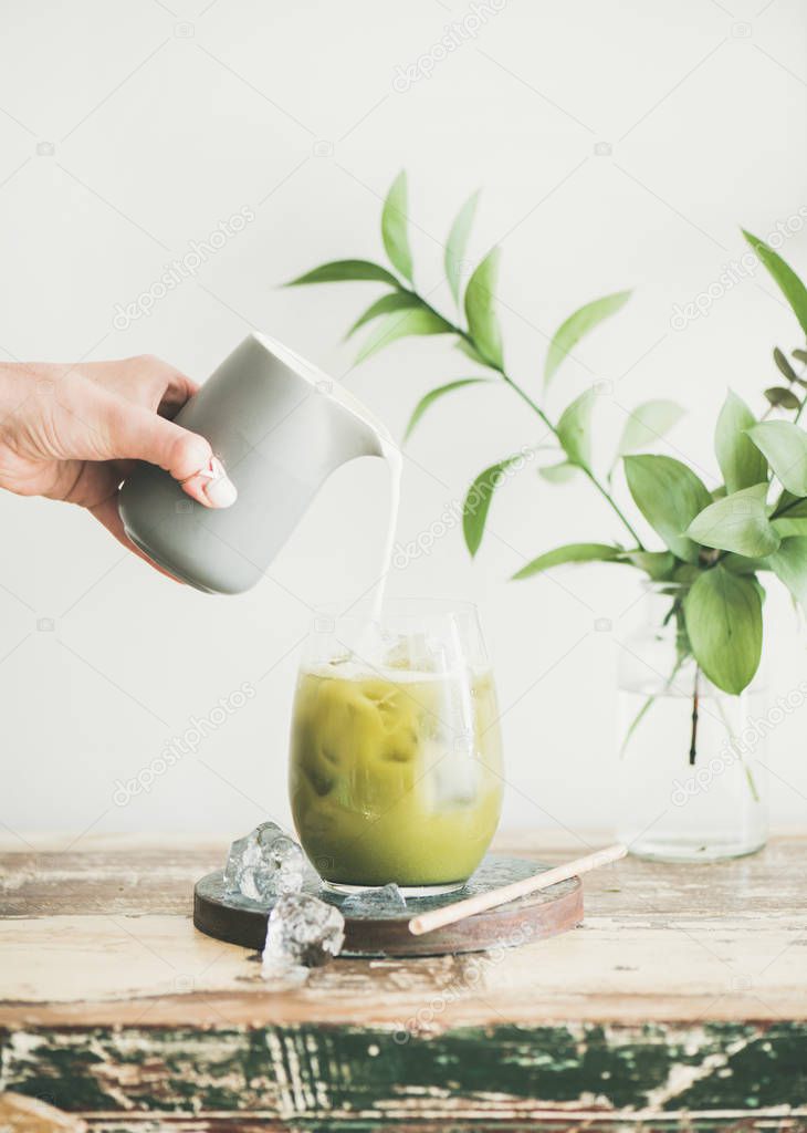 Iced matcha latte drink in tumbler glass on board with coconut milk pouring from pitcher by hand, white wall and plant branches at background