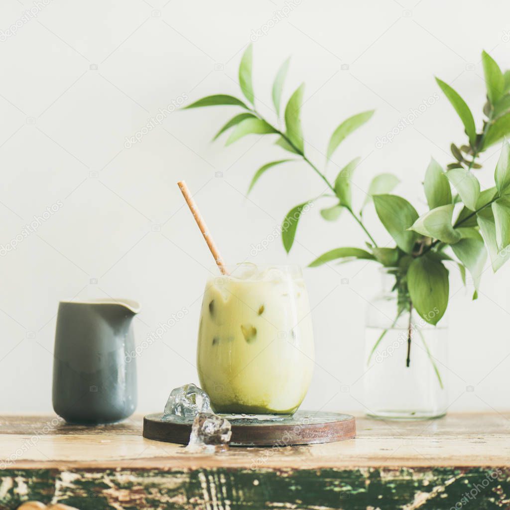 Iced matcha latte drink in glass with coconut milk pouring from pitcher by woman's hand, white wall and plant branches at background