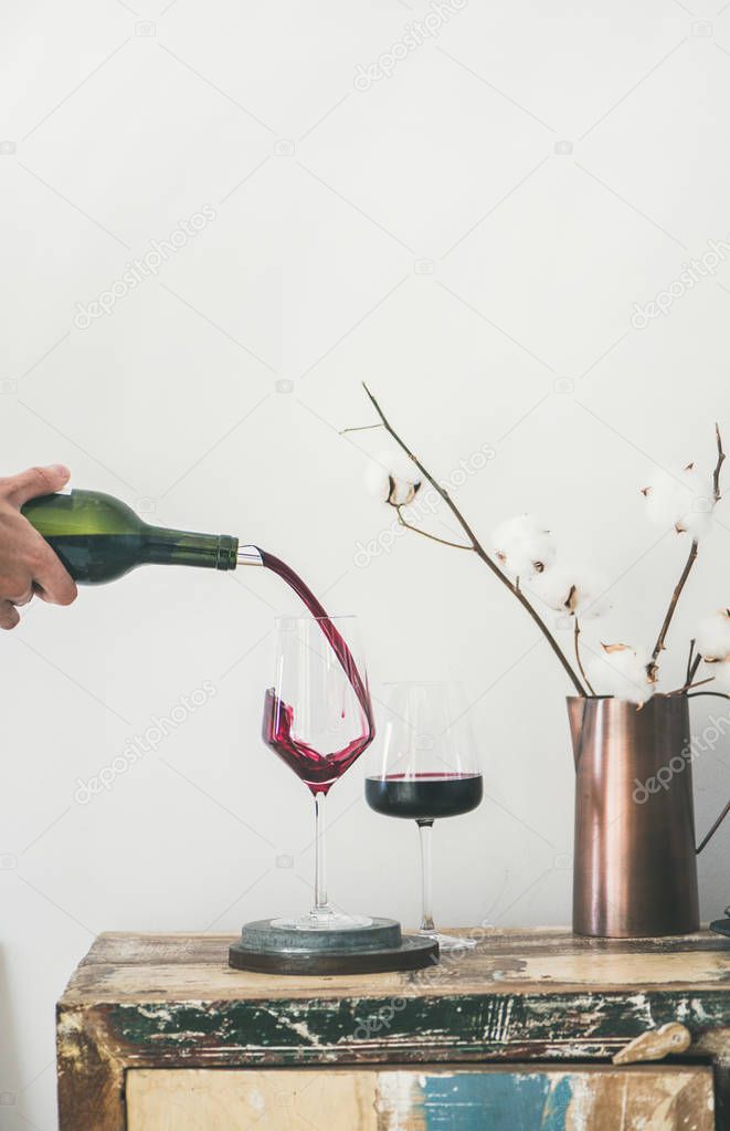 Red wine pouring from bottle into glasses over rustic kitchen countertop, white background behind
