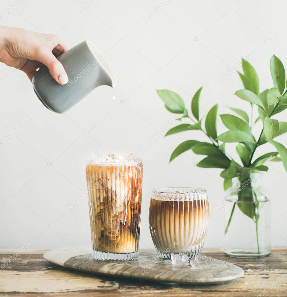 Iced coffee in tall glasses with milk pouring from pitcher by hand, white wall and green plant branches at background