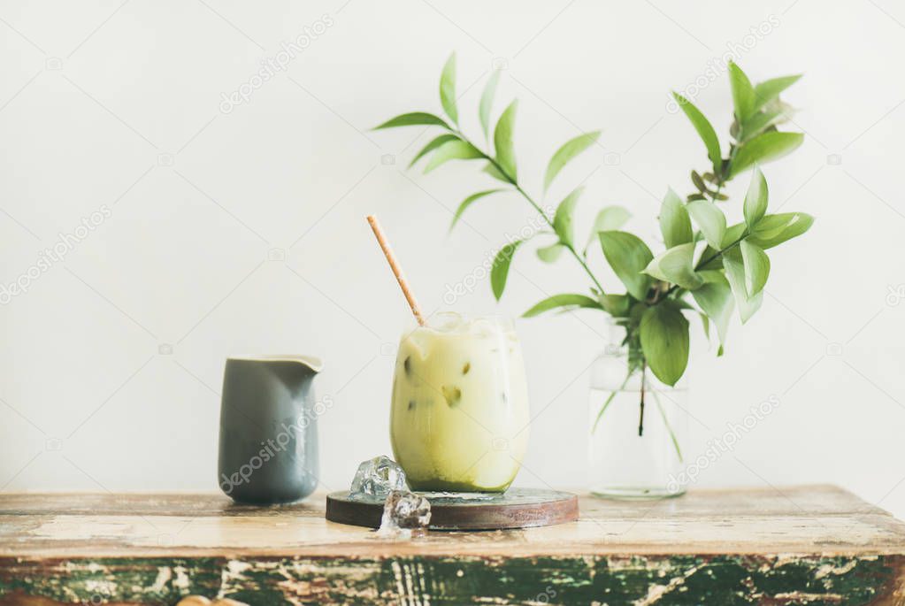 Iced matcha latte drink in glass, white wall and plant branches at background