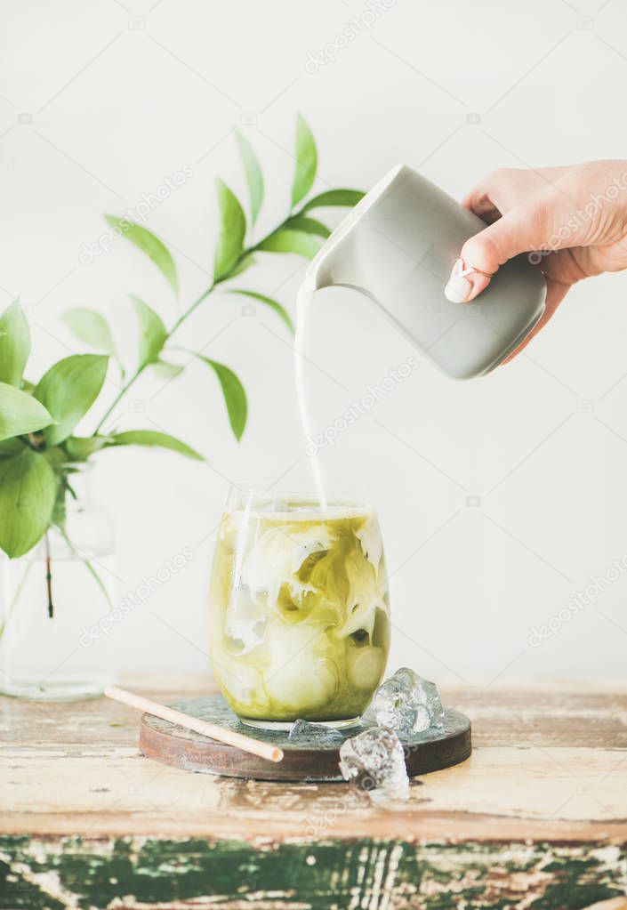 Iced matcha latte drink in glass with coconut milk pouring from pitcher by woman's hand, white wall and plant branches at background