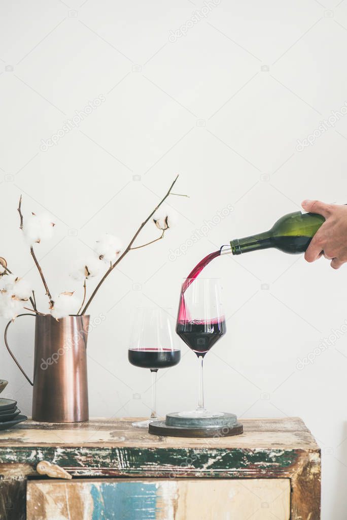 Red wine pouring from bottle into wineglass over rustic kitchen countertop, white background behind