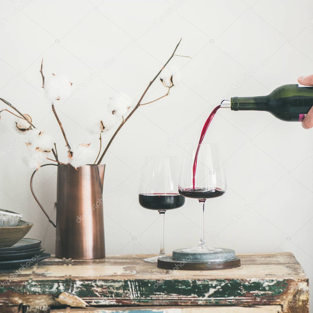 Red wine pouring from bottle in man's hand into wineglass over rustic kitchen countertop, white background behind, copy space, square crop. Wine shop, winery, bojole nouveau holiday concept